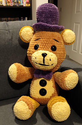A crochet golden yellow bear. He has a light yellow belly, paws, ears and muzzle. He wears a purple hat and bow tie. He has 2 black buttons on his belly.