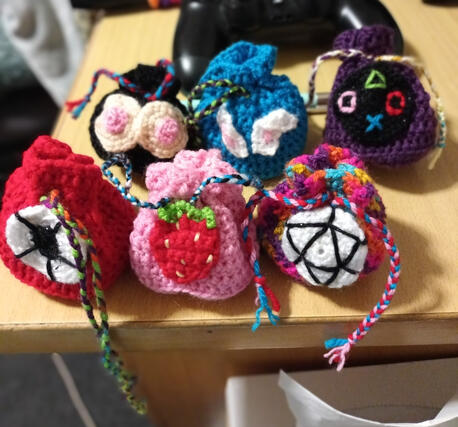 6 small dice bags. A black dice bag with boobs decal, a blue dice bag with bunny ears decal, a purple dice bag with a playstation decal, a red dice bag with a football decal, a pink dice bag with a strawberry decal and a rainbow dice bag with a D20 decal.