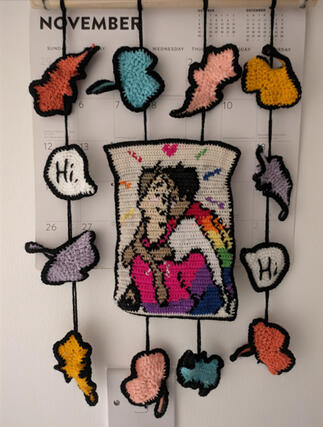 A crocheted wall hanging featuring an image of 2 boys in the centre. One has light skin and blond hair and is wearing a hoodie in the colours of the bisexual flag. The other boy has darker skin and black hair and is wearing a white t-shirt and a cape with